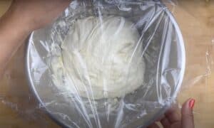 Bread dough rising in a silver bowl covered with plastic wrap, ready for proofing on a wooden surface.